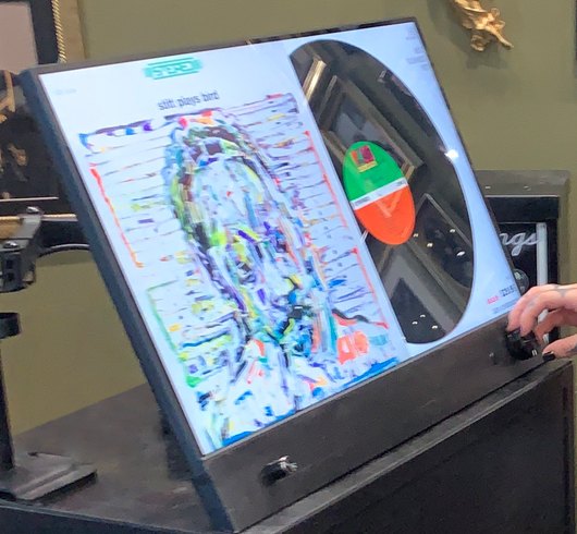 Close view of the Almont display's tilted touchscreen showing a life-size album cover next to its vinyl record and label, with a hand on the control knob.